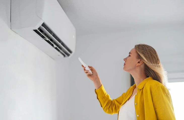 Femme en chemisier jaune utilisant une télécommande pour contrôler un climatiseur mural dans un intérieur moderne aux murs blancs
