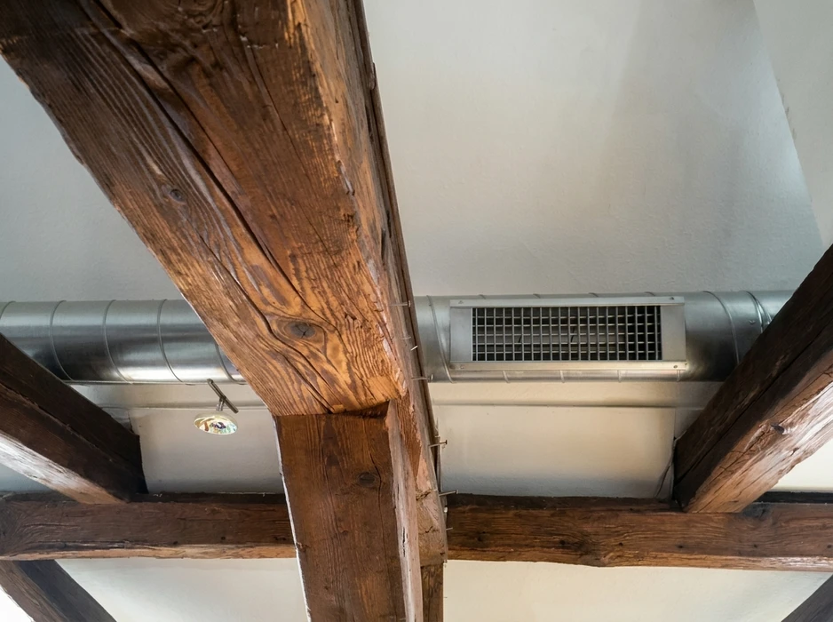 Plafond intérieur rustique photographié en contre-plongée, mêlant poutres apparentes en bois massif ancien et installations techniques modernes