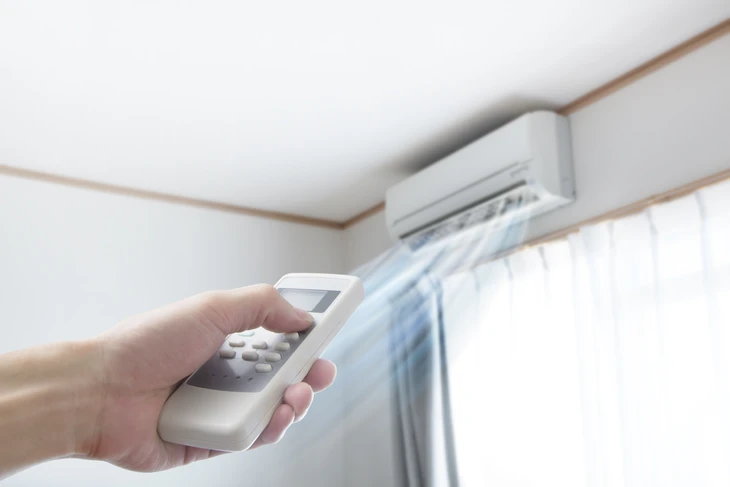 Main tenant une télécommande blanche pointée vers un climatiseur mural dans une pièce moderne avec fenêtres et rideaux clairs