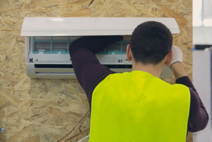 Intérieur d'habitation en construction, technicien en gilet haute visibilité jaune effectuant maintenance ou installation d'unité de climatisation murale