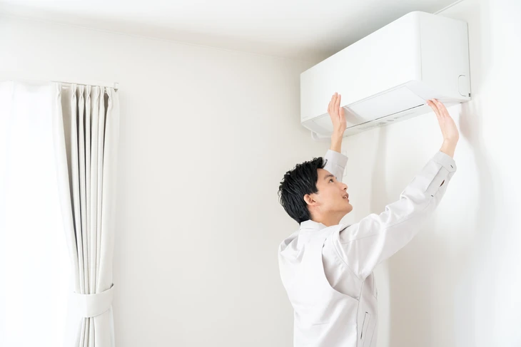 Technicien en uniforme blanc effectuant la maintenance d'un climatiseur mural split dans un intérieur résidentiel moderne aux murs blancs avec rideaux gris