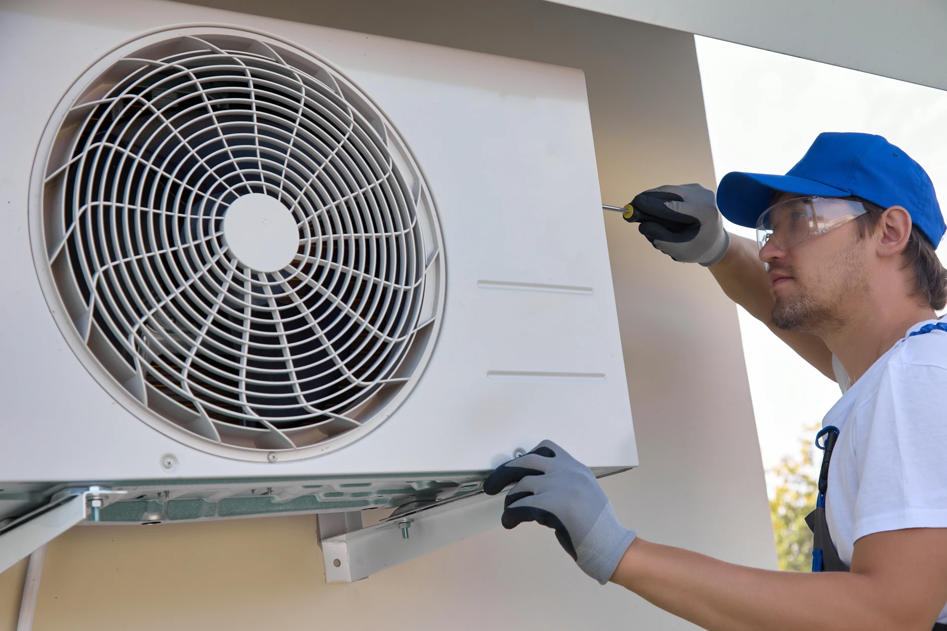Technicien en casquette bleue et polo blanc effectue la maintenance d'une unité de climatisation murale dans un bureau aux plafonds blancs